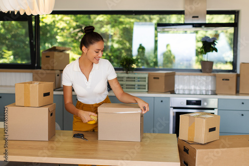 Konstfotografi Smiling young woman taping cardboard box while unpacking in modern kitchen