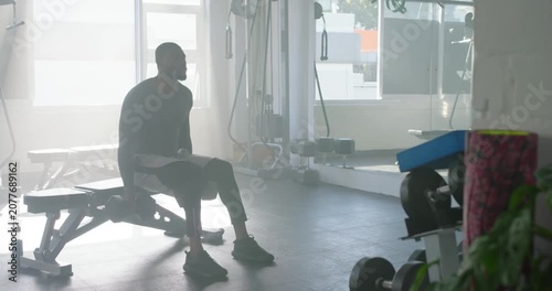 African American man in tee sitting on bench watching light reaching for weight, curling for power