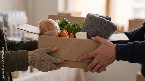 A person hands a cardboard box filled with food and a blanket to another person in a room.