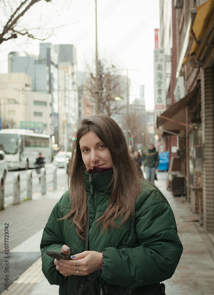 Fototapeta premium Young woman in green puffer jacket holding smartphone smiling on a Tokyo street with traditional shop signage and city buildings in background 