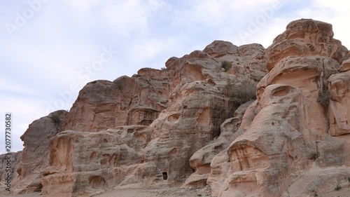 A scenic view of weathered sandstone cliffs and ancient rock carvings in the historic site of Little Petra, Jordan, under a cloudy sky.