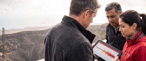 Scientists analyzing seismic data tablet outdoors looking closely at earthquake waves during geological field research project monitoring natural volcanic activity in remote mountain canyon valley.