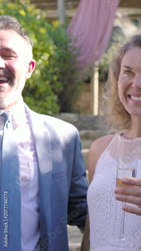 Vertical video: Toasting couple in gray suit, bride raising flutes in yard as guests join wedding