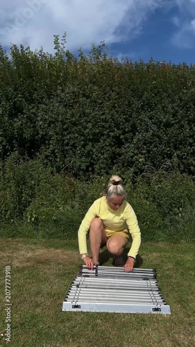 Senior female tourist unrolling and unfolding a portable aluminum camping table on green grass.