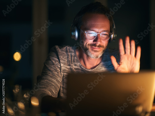 A man wearing headphones and glasses smiles warmly while waving to a laptop screen, signifying a video call or online meeting in the evening.