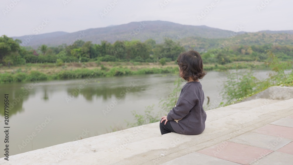 Fototapeta premium A young toddler sits alone on a concrete embankment, quietly observing the river and mountains, embodying a peaceful moment of childhood contemplation.