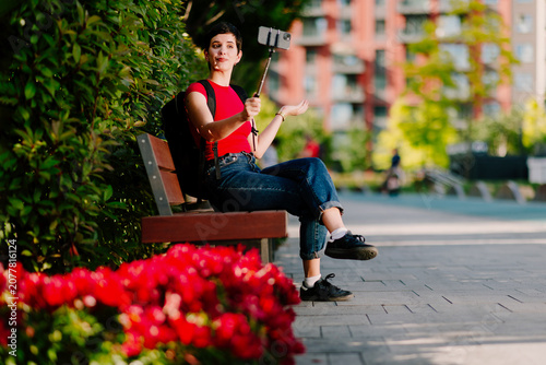 Young Caucasian woman sitting on a park bench while recording online content with a smartphone mounted on a selfie stick.