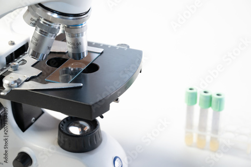 Close-up of laboratory microscope with sample glass slide and blurred blood test tubes on white background, copy space.