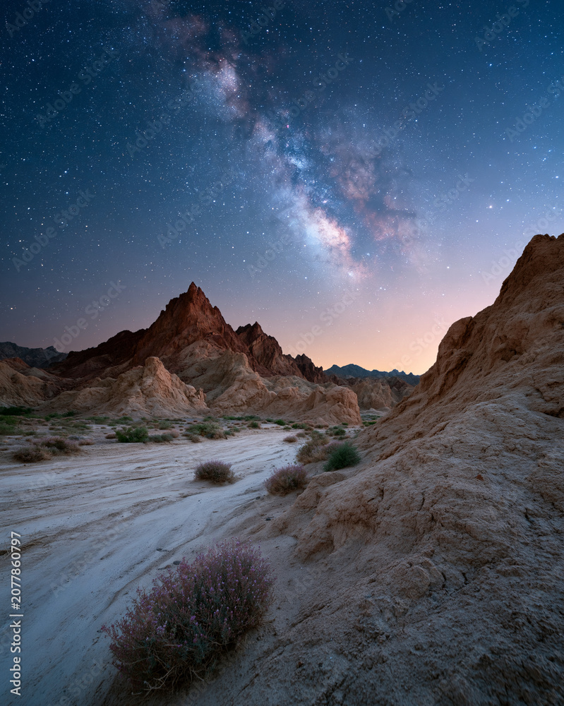 Obraz premium Milky Way arches above rugged desert peaks. Soft twilight paints sky with hues of rose and blue. Distant stars glitter like scattered diamonds. Dry earth and sparse shrubs frame the scene