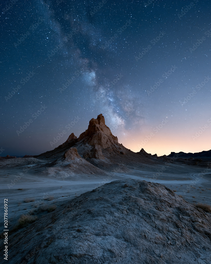 Fototapeta premium Dark desert peak rises beneath swirling Milky Way. Starry sky glows softly above rocky horizon. Dusk paints horizon with warm, fading light. Silent, ancient land holds timeless cosmic wonder