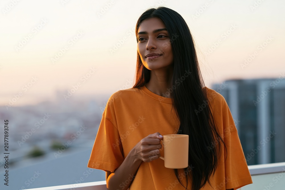 Fototapeta premium Young woman on rooftop balcony holding coffee mug at sunrise with soft city view for lifestyle background and wellness morning routine design