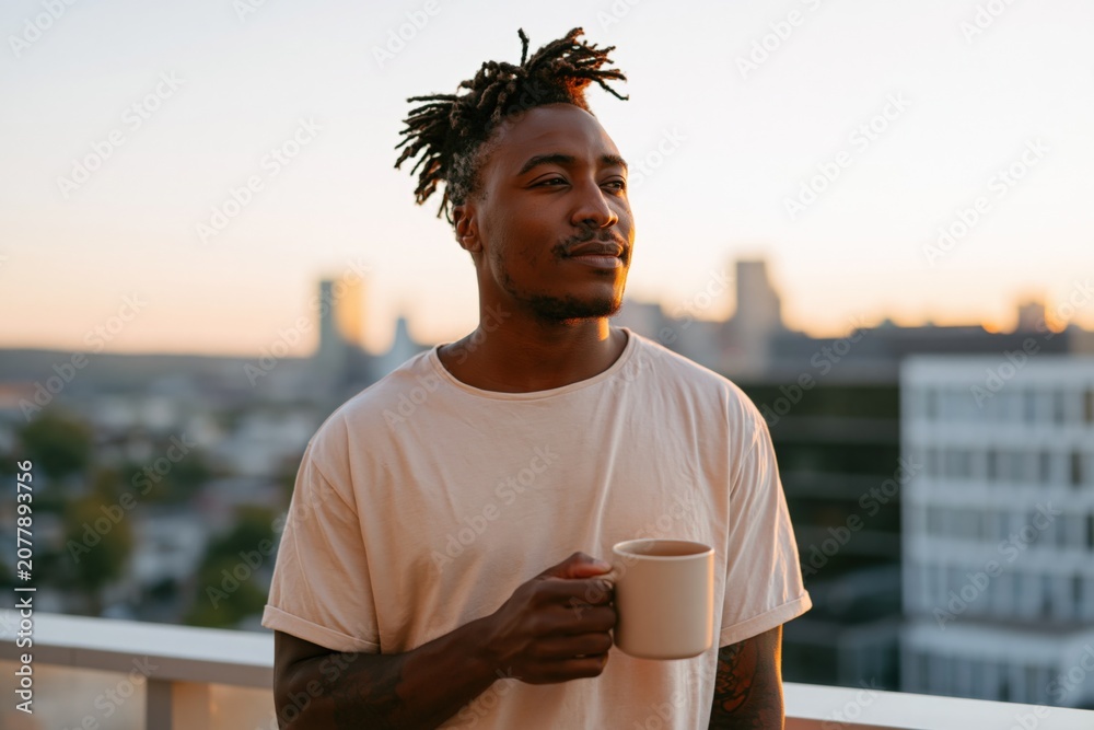 Fototapeta premium Young man holding a mug on rooftop at sunrise with urban skyline for lifestyle banner and peaceful morning background