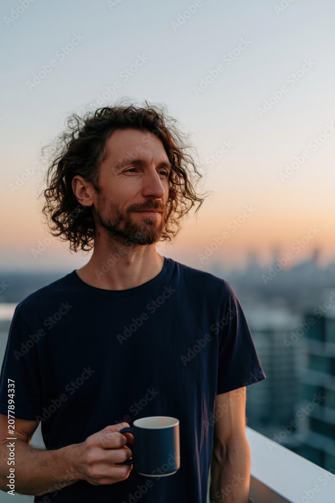 Fototapeta premium Young man with curly hair holding a dark mug on a rooftop balcony at sunset for lifestyle portrait and morning routine background for wellness and relaxation banner
