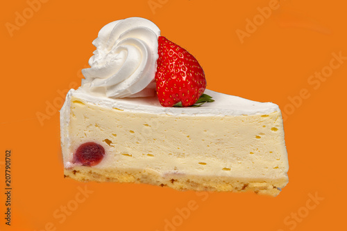 A slice of cream cheesecake is on a plate. It sits on a shortbread base and is topped with fresh strawberries and whipped cream. The background color is bright.