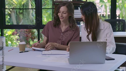 Two diverse businesswomen discussing financial report using laptop and calculator at modern office desk. Smiling and collaborating on successful project.