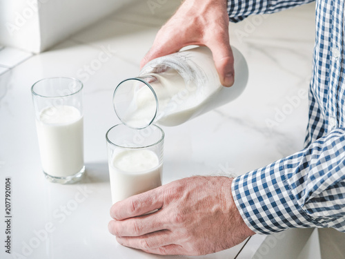Fotografie Man's hand pours fresh milk from a glass bottle into a clear glass on a white marble table