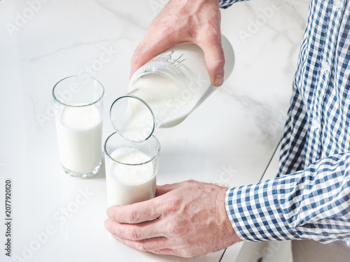 Fotografie Man's hand pours fresh milk from a glass bottle into a clear glass on a white marble table