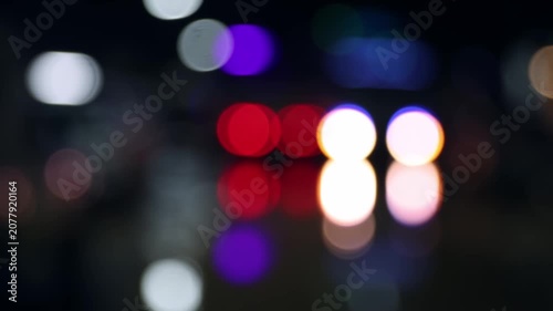 Defocused night traffic light on wet street after rain. Beautiful glowing colorful bokeh effect and abstract urban background with reflection.