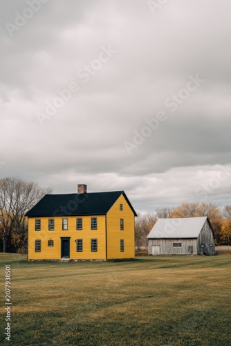 Bright yellow house stands next to a simple gray barn in an open field under a cloudy sky during autumn in a rural setting