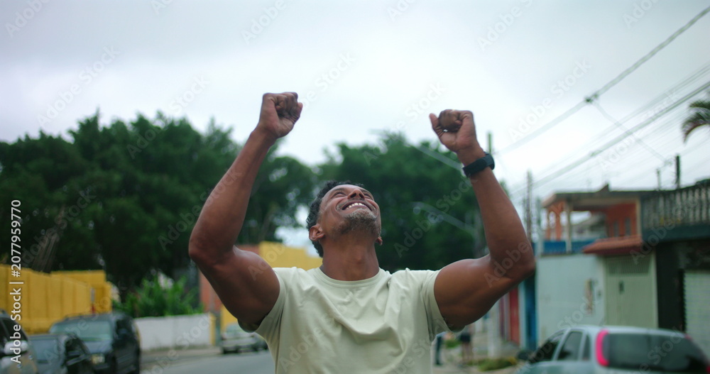 Obraz premium Man of African descent standing on urban street with arms raised, looking up in gratitude, emotional moment of relief and hope, expressive gesture of thankfulness and faith