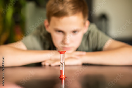A thoughtful young teenager kid boy resting her head on a table next to a hourglass