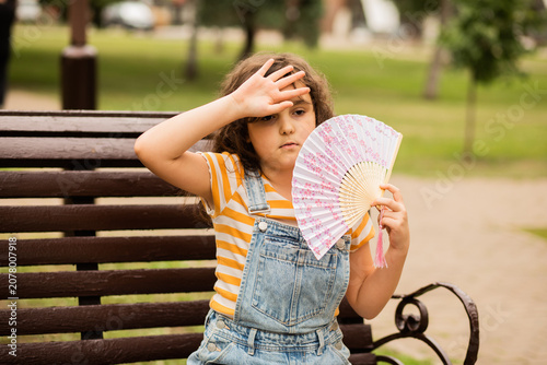 Canvas Print Cute little girl kid child is sitting on a park bench, fanning herself with a decorative fan to cool off