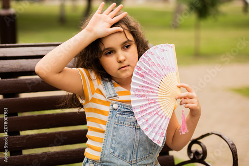 Fotografija Cute little girl kid child is sitting on a park bench, fanning herself with a decorative fan to cool off