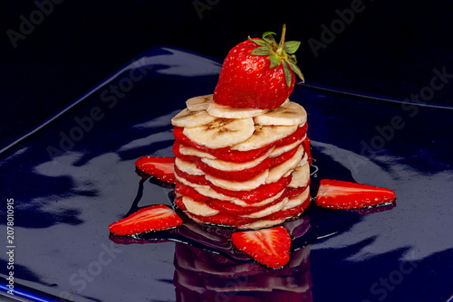 This dessert tower is made of sliced bananas and strawberries. It features a whole strawberry on top and is decorated with honey. It is set on a shiny blue plate.