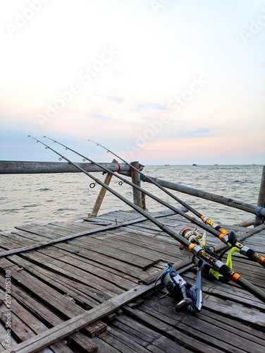 Fishing rods resting on a rustic bamboo pier overlooking the calm sea at sunset, creating a peaceful coastal fishing scene with a tranquil evening atmosphere