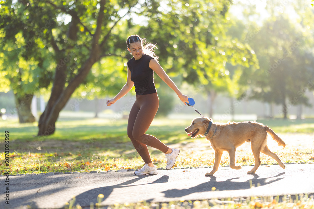 Obraz premium Young woman walking a golden retriever dog on a sunny day in a park, wearing athletic clothing and enjoying outdoor exercise along a paved path