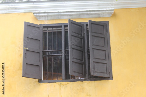 Old brown wooden window frame with open shutters on yellow plastered exterior wall.