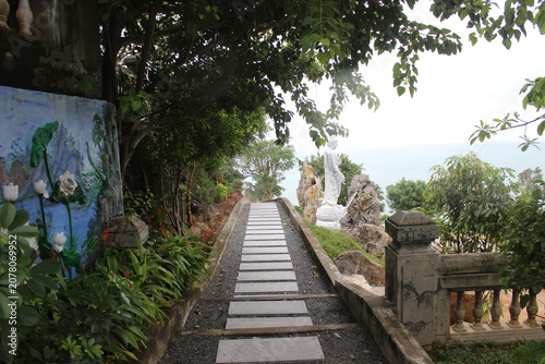 Stone pathway leading to white statue overlooking calm ocean in tropical landscape.
