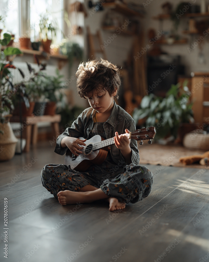 Fototapeta premium Young boy playing ukulele while sitting on wooden floor indoors. Child sitting on floor with ukulele