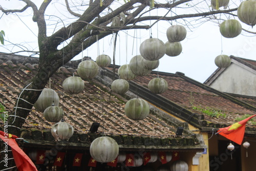 Neutral colored paper lanterns hanging from branches above traditional roof tiles.