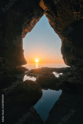 Rocky cave opening frames a calm sunrise over the sea with reflections in tidal pools on the Sardinian coast.
