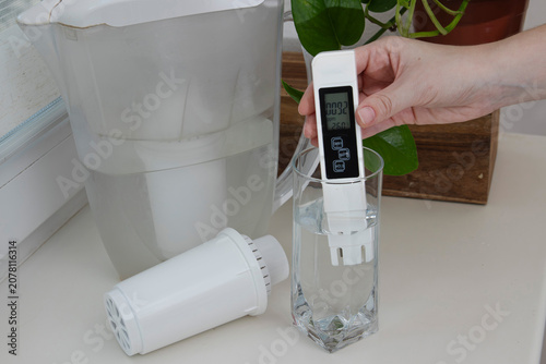 Hand measuring filtered drinking water with a digital TDS meter beside a household filtration pitcher and replacement cartridge. Concept of water purification, clean drinking water, water quality test