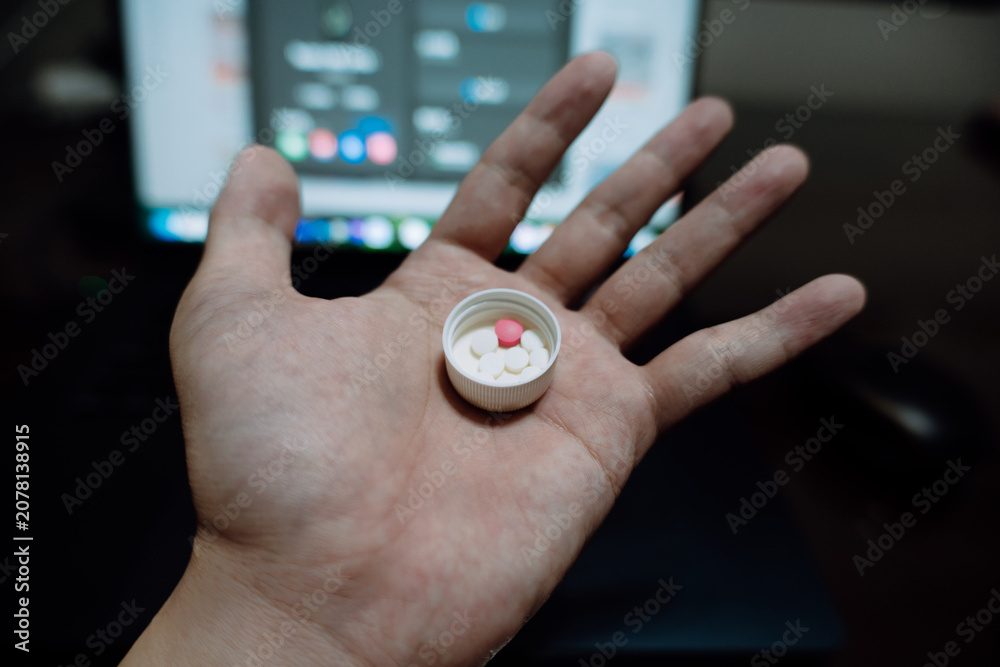 Fototapeta premium Hand holding small white pill cap with red tablet inside, blurred laptop background — close-up
