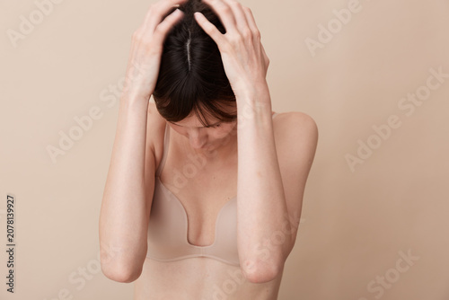 Obraz na plátně Young woman in a beige bra stands against a soft neutral background, hands in her hair, conveying subtle tension and vulnerability in a quiet moment