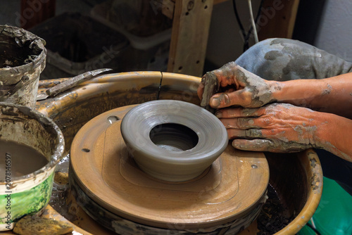 Clay covered hands shaping and trimming a wet clay vessel on a spinning pottery wheel in a ceramic studio. Selective focus. Motion effect.