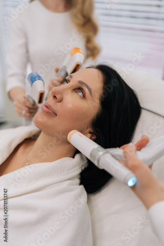 Close-up vertical portrait of woman receiving various professional aesthetic treatments including ultrasound and advanced devices by cosmetologists for skin care and rejuvenation in beauty clinic.
