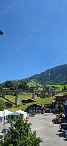 A high-angle vertical shot of a traditional Swiss alpine village with wooden chalets scattered across green rolling hills, featuring a large concrete viaduct bridge stretching across the valley under 