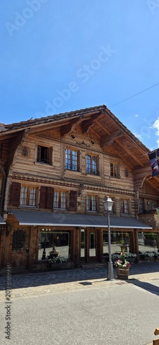 A low-angle vertical shot of a classic alpine wooden chalet style building featuring traditional window shutters and a ground-floor storefront under a clear blue summer sky in Switzerland.
Category