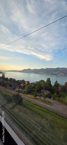 A stunning high-angle shot captured from a train window showing the beautiful Lake Thun surrounded by traditional houses, green hills, and distant mountains under a sunset sky in Switzerland.