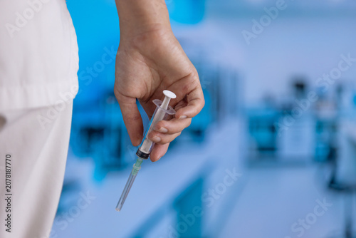 Hand grips a syringe. The syringe has a needle and plunger. Blurred background shows a medical lab setting. White scrubs are visible on the arm. This image captures healthcare professional work