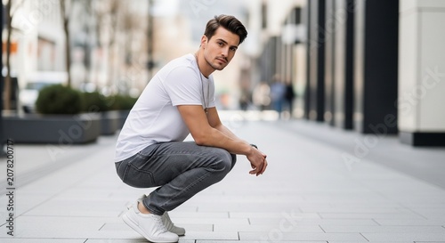A stylish young man in a white t-shirt and grey trousers squats down on a clean city pavement, looking directly into the camera with a confident expression