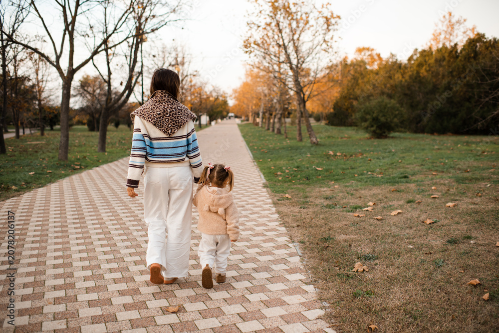 Fototapeta premium Happy mother with kid girl daughter walking in autumn park wearing knitted clothes over yellow leaves rear view. Fall season. Motherhood.