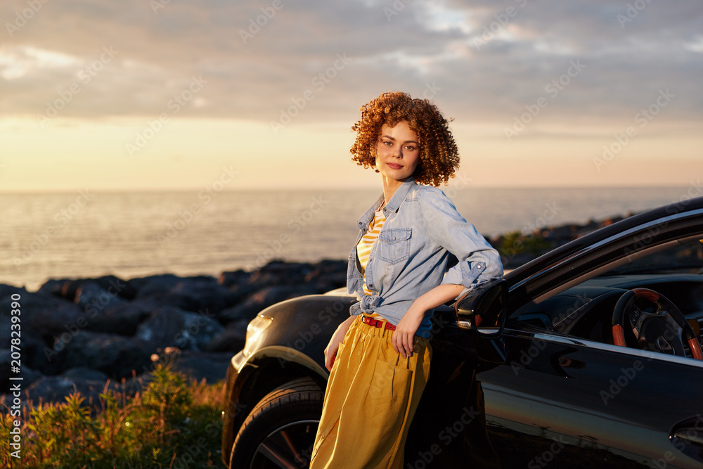 Fototapeta premium A stylish woman stands beside a parked car at golden hour by the sea, wearing a denim jacket and yellow dress. A lifestyle portrait conveying confidence and outdoor freedom.