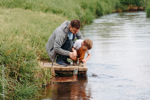 Dad and son launch paper boat on small river, young caucasian boy with adult man, father`s day, sunny summer day, green grass, children games and fun