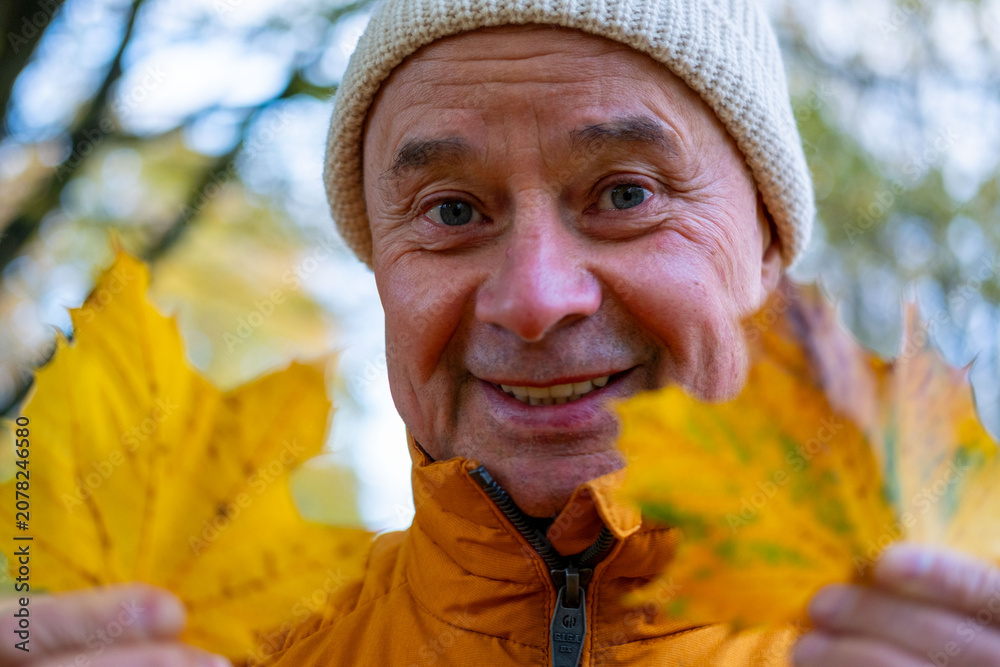 Naklejka premium Smiling senior man in orange jacket holds bright yellow autumn leaves, looking joyfully at camera, surrounded by soft fall foliage, Joyful senior man celebrating autumn’s beauty, seasonal happiness