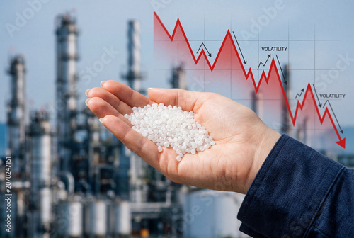 Hand holding white plastic pellets against a refinery background with a downward volatility graph